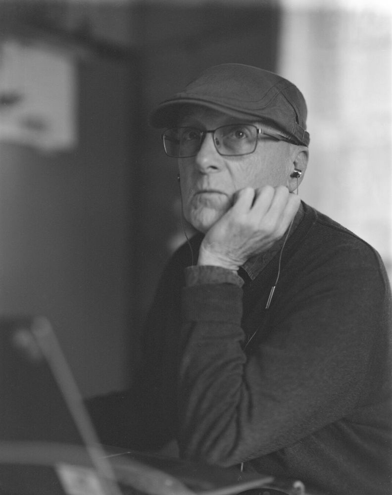 A middle aged man with wire framed glasses, a hunting cap, and earphones in his ears looks off into the distance while leaning his had on his hand.