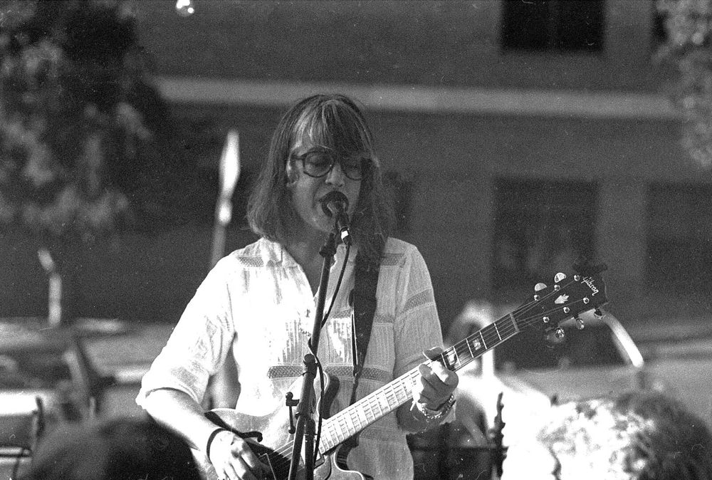A middle aged woman with graying hair cut into bangs plays guitar and sings into a microphone.