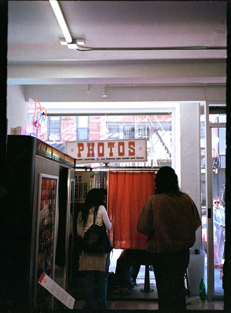 Two people stand in front of a photobooth, facing away from the camera. The photobooth is in the window of a storefront, and has a sign on top saying “* Photos * Electronic Lighting * All photos are sharp and clear”. Another photo booth is perpendicular to it against a wall and has a sign that says “Color Photos”.
