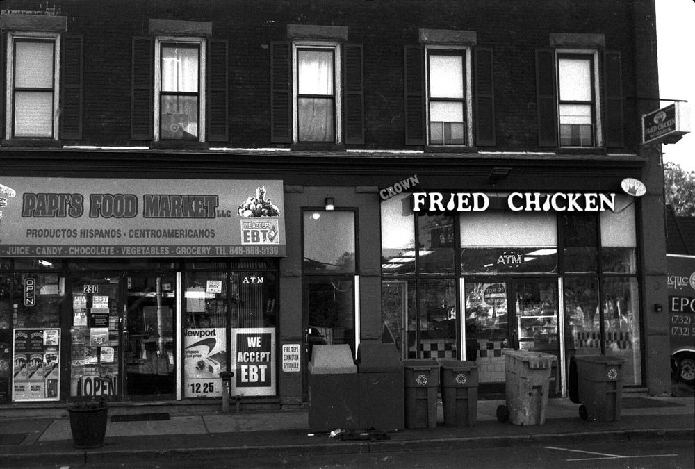 A small grocery store and a fried chicken restaurant stand next to each other. The grocery on left has an awning that says “Papi’s Food Market LLC Produtos Hispanos • Centroamericanos” with a cornucopia next to it. A sign added to the sign says “We accept EBT”. Below all that is a list of product types: “Juice • Candy • Chocolate • Vegetables • Grocery Tel 848-868-5130”. There is an OPEN sign in the window, and an ad for Newport cigarettes in another window. To its right, Crown Fried Chicken with a sign where each I in “FrIed ChIcken” is replaced with a drumstick. A sign reading “ATM” is in the window above the door. On the door is a backward reading reflection of a street sign that says “Do Not Enter”.