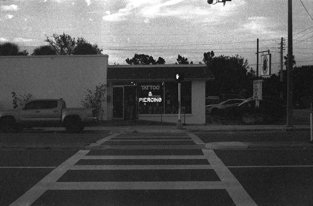 A one story building with a glass door and windows with a sign in the window that says, in neon, “Tattoo & Piercing”.