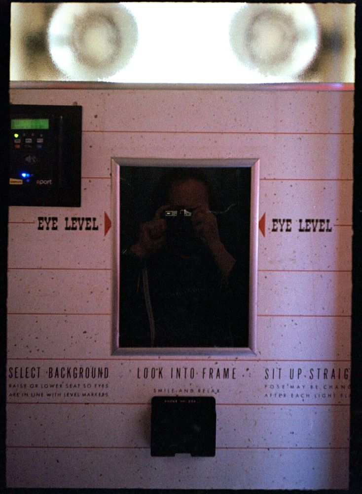 The view from the seat of a photo booth. Two flashes are behind frosted glass at the top, looking like eyes. The glass covering the camera looks like a nose. Something below that looks like a mouth.  The words “Eye Level” appear next to the camera, with arrows pointing toward the camera. Below the camera, instructions: “Select Background. Raise or Lower Seat So Eyes Are In Line With Level Markers. Look Into Frame. Smile and Relax. Sit Up Straight. Pose May Be Changed After Each Light Flash.”
