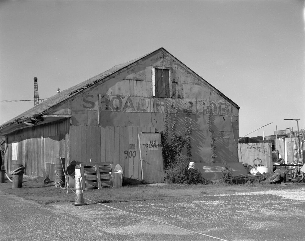 A run down wooden building sits on a lot. A sign painted on the eaves says “Shoal Harbor”. A door in the center of the wall is off its hinges. The words “No Trespassing” are painted on the door.