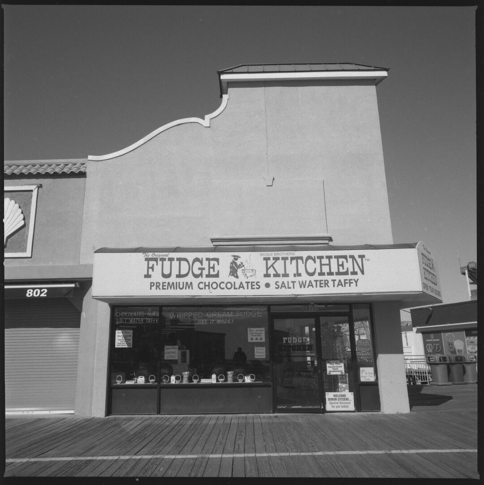 A candy stand on the boardwalk. The building is stucco and the stand is at the corner. The corner of the building has a tower. The stand has a full wall of windows at ground level. An awning over the front reads “The Original Bogle Bros. Fudge Kitchen. Premium Chocolates • Salt Water Taffy”.