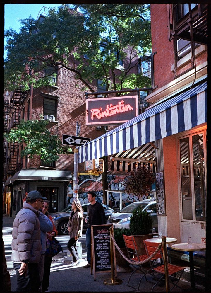 People stroll past a restaurant. A chalkboard outside lists specials. A neon sign standing out from over the door reads “Rintintin”.