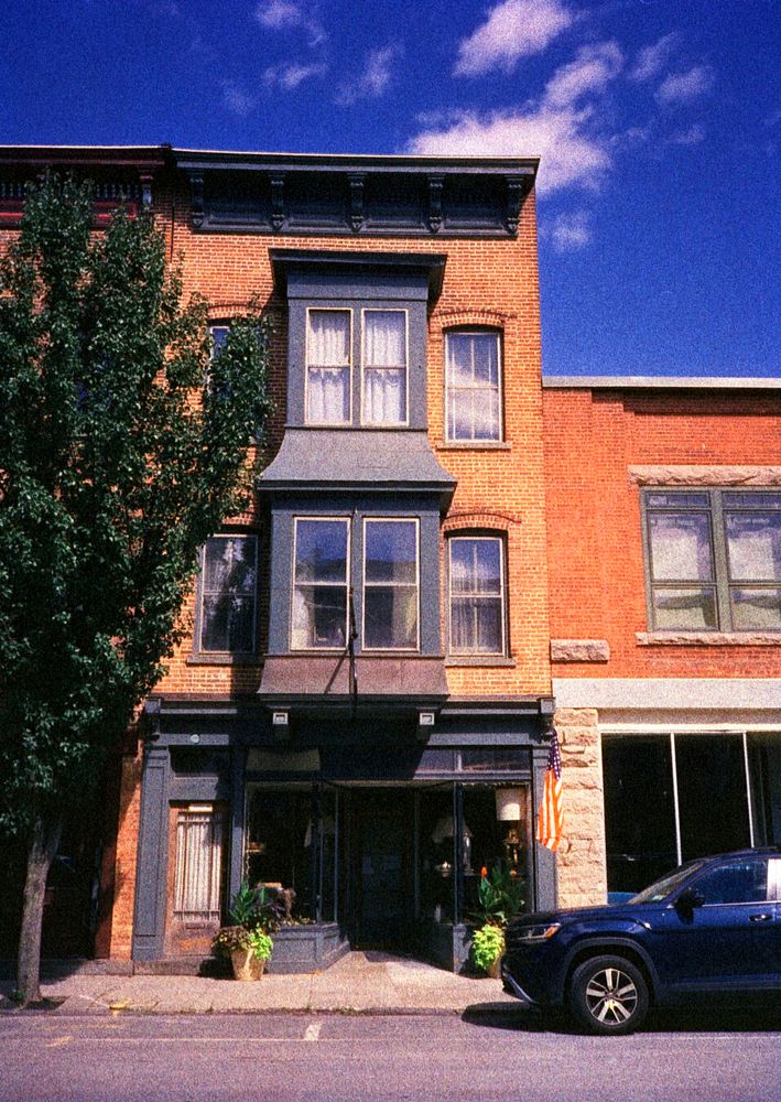 A three story brick building stands close to the street. The ground floor houses a store.