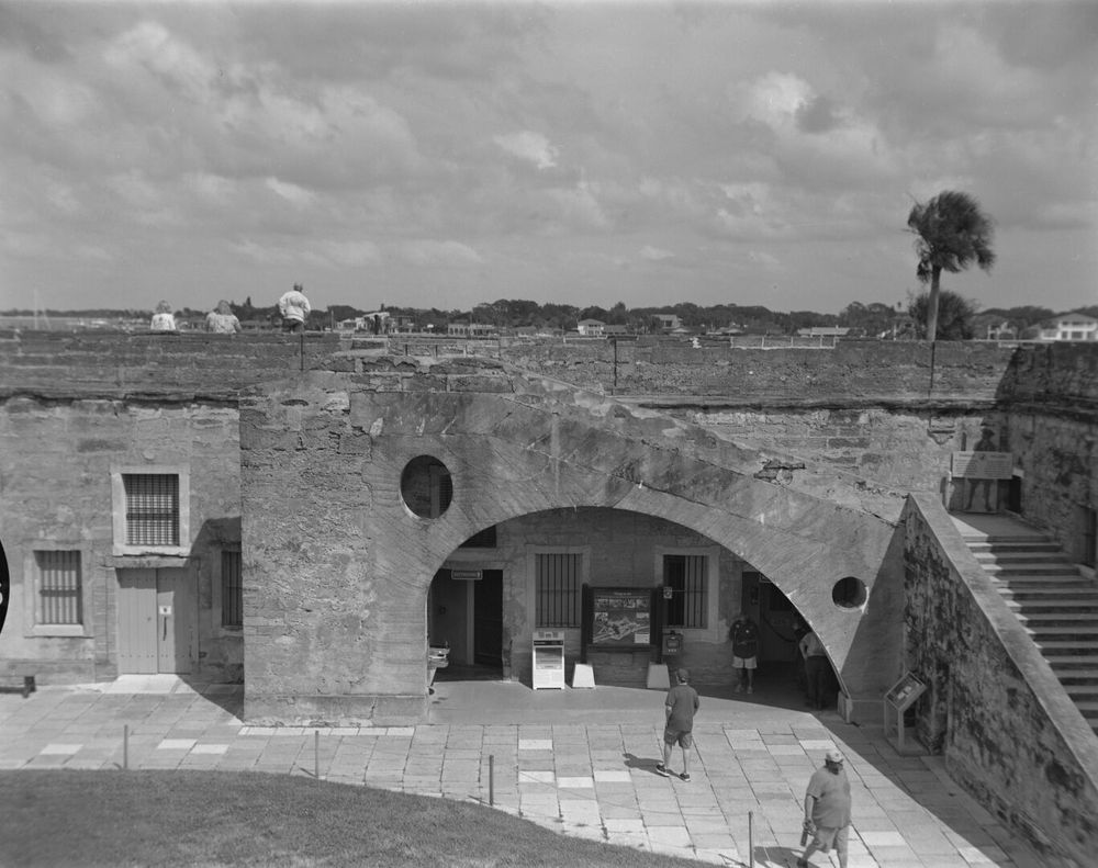 A few people wander around the courtyard of a stone fort. A few others stand on the wall. The city is visible across the water.