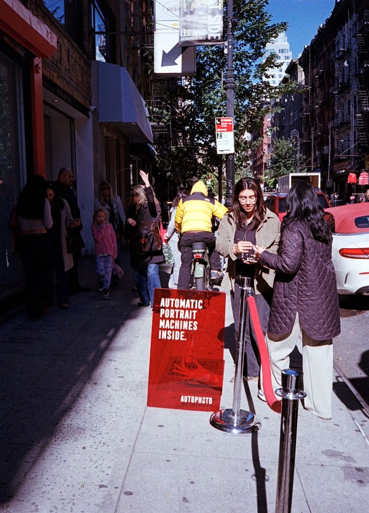 A transparent red sign on the sidewalk says “Automatic Portrait Machines Inside. Autophoto.” Two long haired people stand next to the sign in sunlight. Other people stand against the building in shade. A red rope barrier is notable for not apparently serving any purpose.