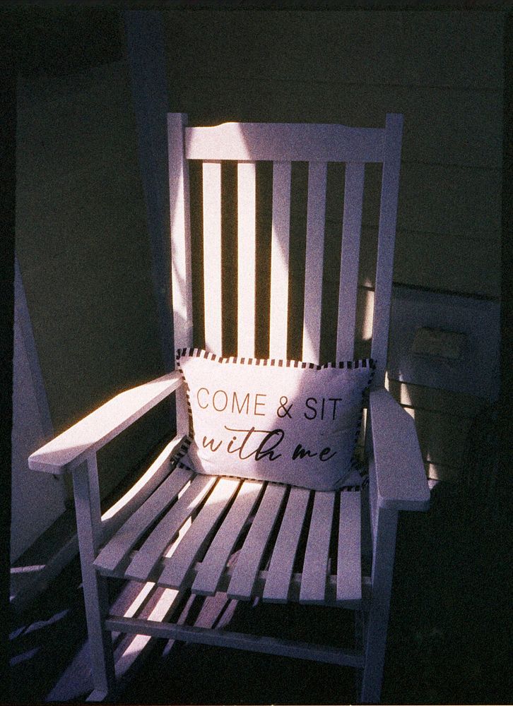 A white rocking chair sits on a porch with a pillow on it that says “Come & Sit With Me”.