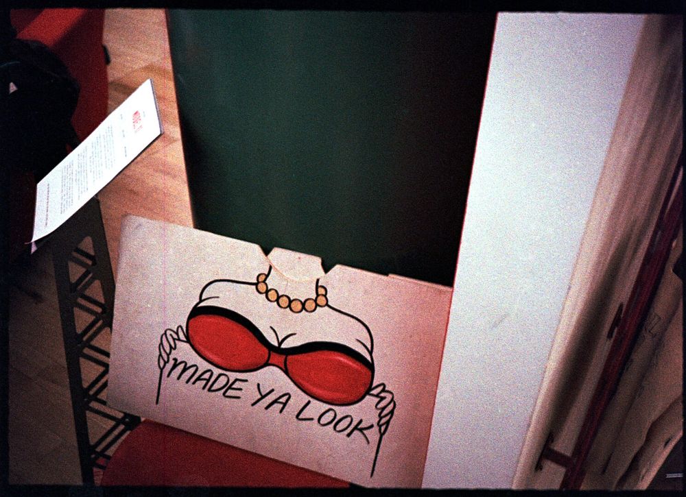 A sign showing a drawing of a woman’s torso with prominent breasts covered by a red bra, pearls around her neck, holding up a sign that says “Made Ya Look”.