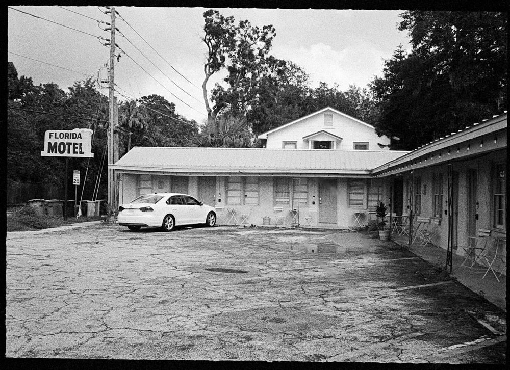 A run down one story motel in an L shape. A sign to its left says it is the Florida Motel. There is one car parked in the parking lot. The top of a house is visible behind the motel, as are trees. A speed limit sign on the street says the speed limit is 20.