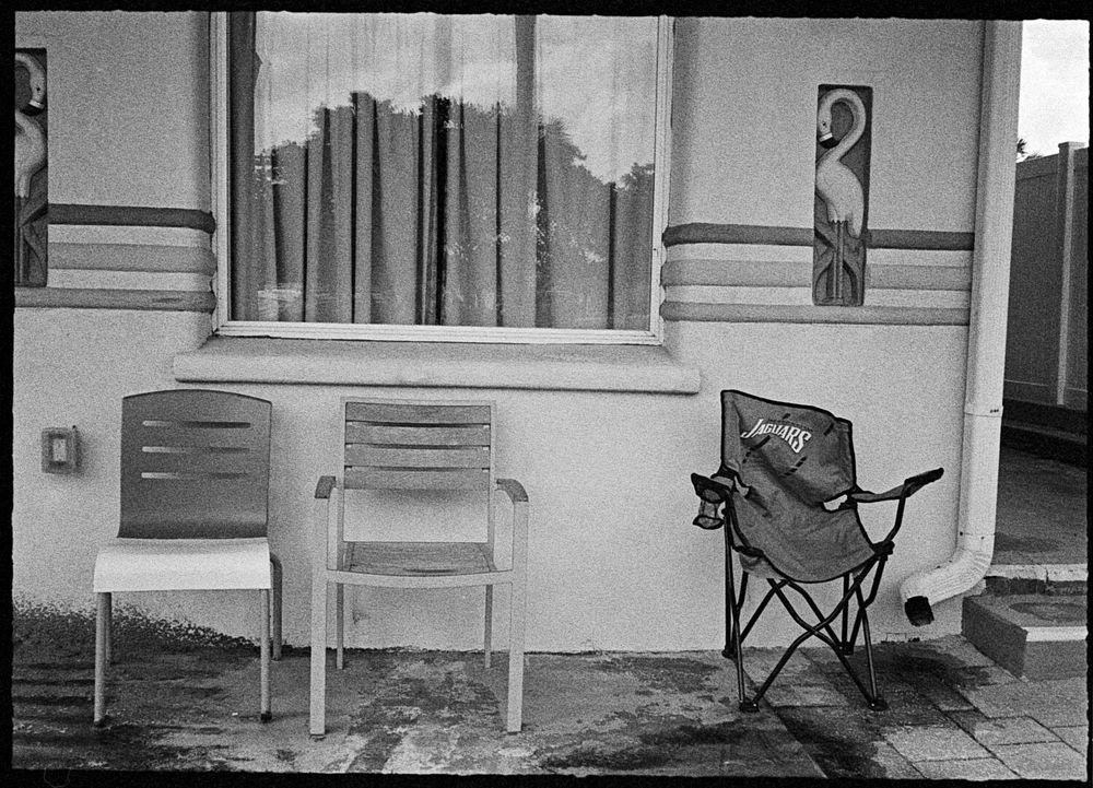 Three chairs sit in front of a motel. One is plastic, one is metal and wood, and the third is a folding carryable chair with the logo of the Jacksonville Jaguars on it. The wall has three colored stripes on it, and flamingos in relief, flanking a room window in the center.
