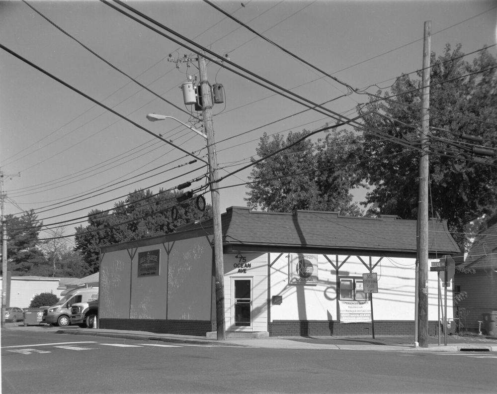 A white stucco building sits at a corner. The entrance is at an angle, and above it is the address, 75 Ocean Ave. On the right, which is sunny, there is a sign for Rheem heating and cooling products and a help wanted sign. On the left in shade is the business sign, All-Phase.