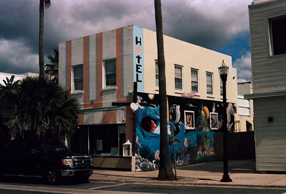 A smallish two story building stands on the street. The front has red stripes with a blue edge on one side painted on the second floor. The first floor is full sized windows. The side of the building has four double pane windows on the second floor, which is painted a beige color. The first floor has an intricate mural painted on it with space travel and a seahorse and other aquatic creatures. There’s also a dog.