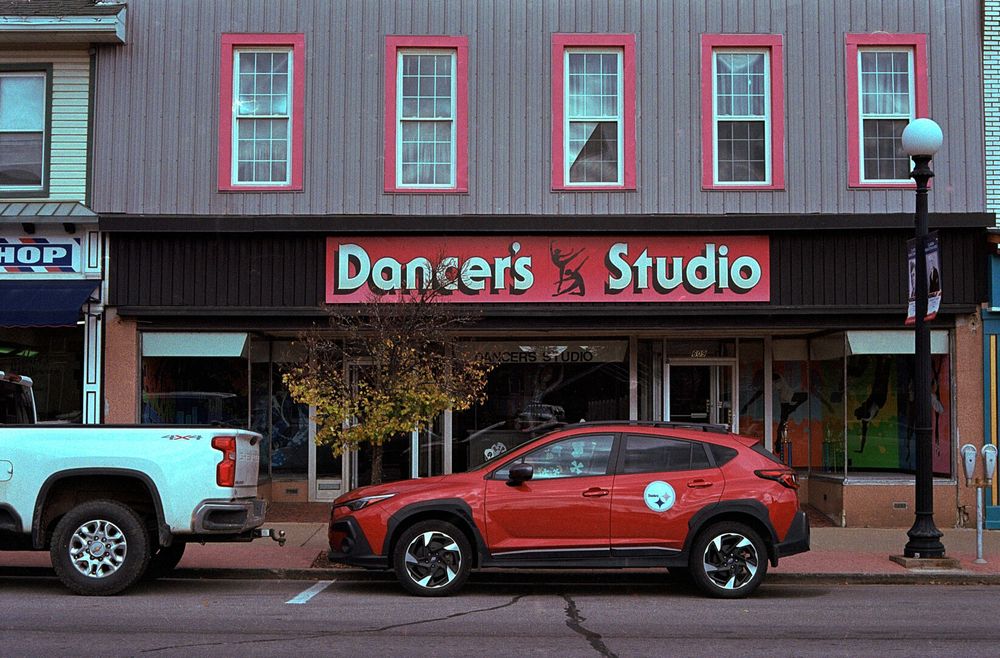 A storefront has a sign above the first floor windows reading “Dancer’s Studio” with two ballet dancers in silhouette between the two words. Supposedly they can’t both fit in the studio at the same time since the studio is singular. There is a second floor with several red framed windows.
