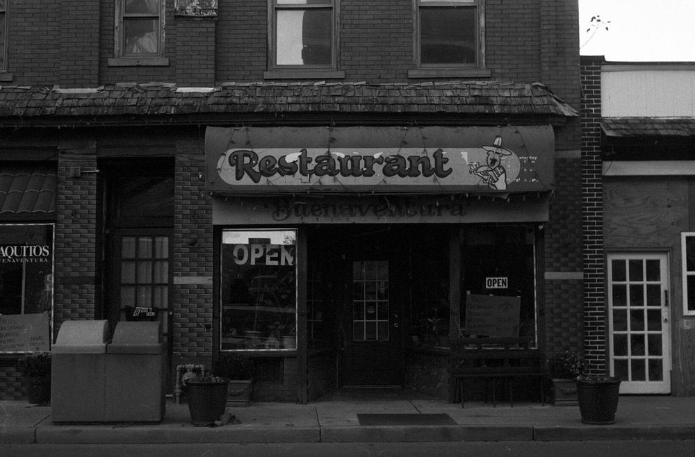 A two story brick building has a restaurant on the first floor. The awning over the restaurant says “Restaurant” with a man in a sombrero and traditional multicolored cloth next to it, with a list of hours superimposed that is largely unreadable.  Below the awning, another sign says “Buenaventura”. Two signs in the windows say “Open”.