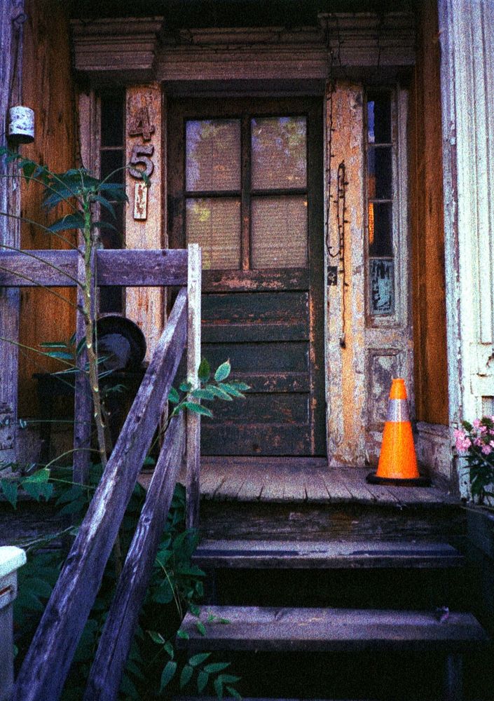 A run down house with worn wooden stairs and railings leading to a small porch, also worn. A traffic cone sits on the porch. The address, 45, is in large numbers to the left of the door. Paint is peeling off the walls, and the door is worn.