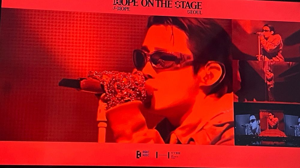Jhope on stage in multiview, close view of his hands on the mic and his head in red light. He’s wearing shades and sparkling gloves 