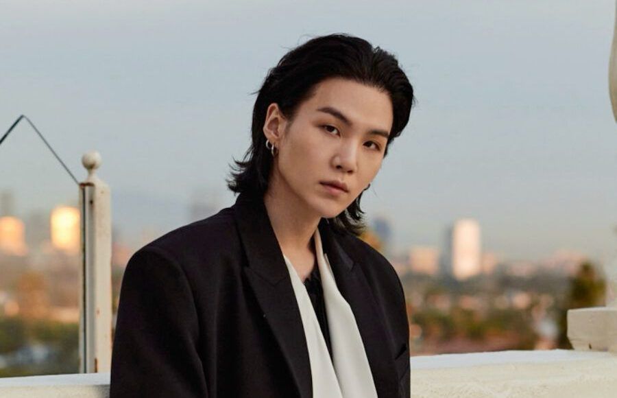 Bts suga portrait picture on a rooftop at sunset, he‘s got long black hair tugged behind the ears, head slightly tilted and looking directly to the Camera. His gaze is piercing and his jawline Sharp. He’s wearing a black suit jacket with a white scarf and black tee. 