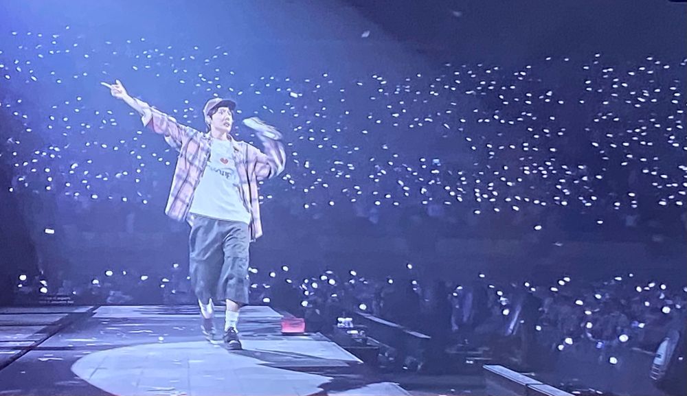 Jhope from bts on Stage in Front of a big crowd, wearing a shirt, cap, sneakers and a white Shirt. 