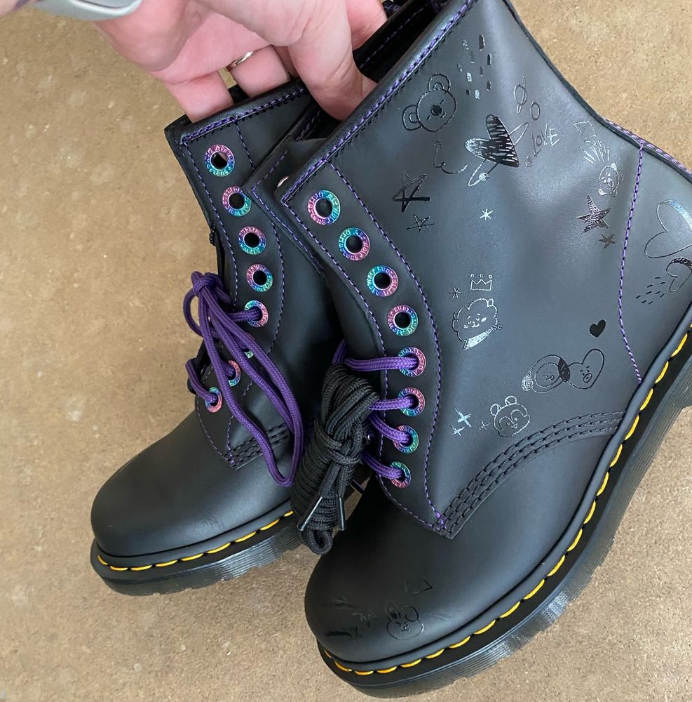 A picture  of  the new  black BT21 collab doctor Martens Boots with purple shoe laces and rainbow Details Held in one Hand. There Are small bt21 Charakters on the side of the boot in Shiny black