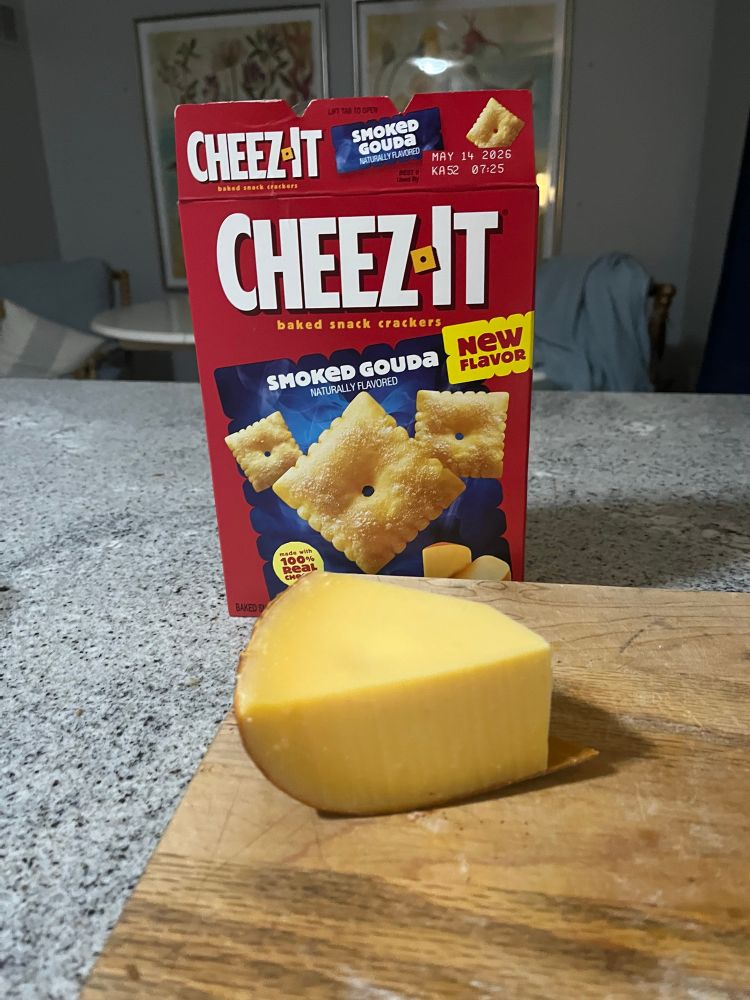 A piece of (not particularly fancy) aged Gouda on a cutting board with a box of smoke Gouda cheezits in the background.
