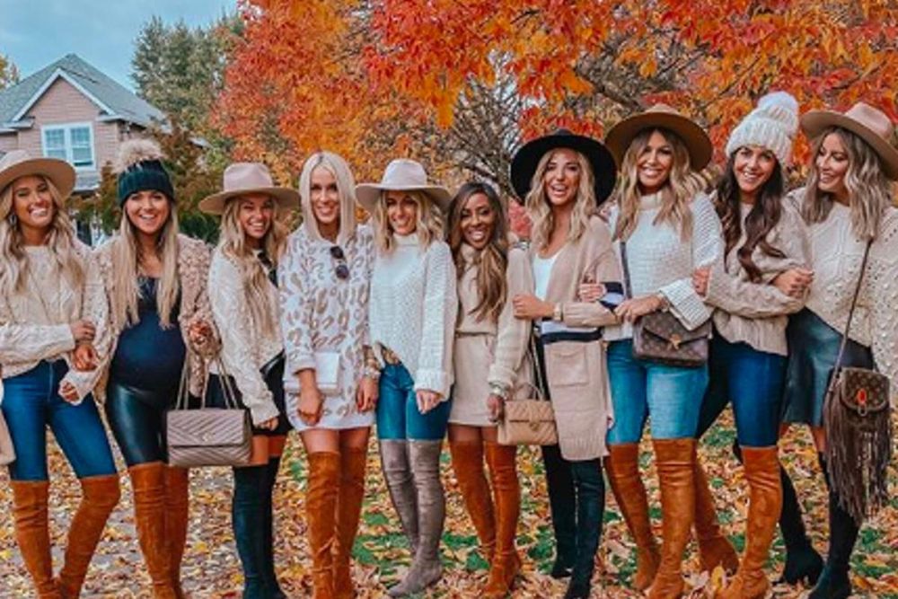 Ten young women posed in a colorful fall outdoor setting. All are wearing very tall boots and blue jeans. Most are wearing broad brimmed Stetsons or stocking caps with big fuzzy poms. They have fabulous, colored and styled hair and gleaming white smiles. 