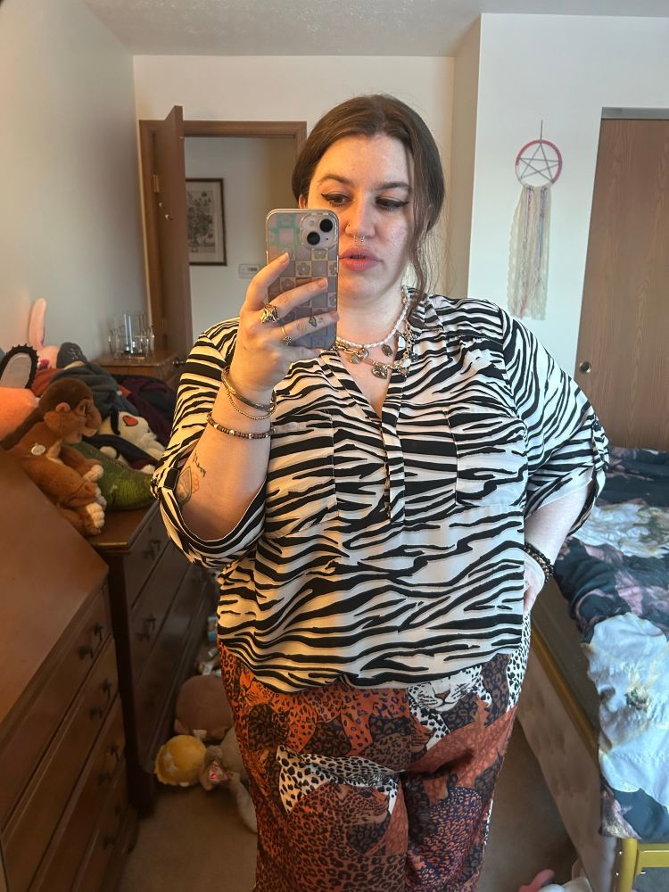Selfie of me wearing pants of various colored leopards printed on them, wearing a blouse of white tiger stripes (some may say zebra stripe but I like white tigers), some bracelets on my wrists, hair up except for a little wave hanging beside my face