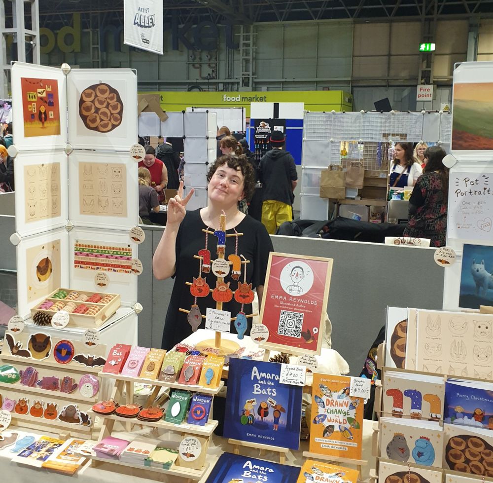 Me Emma Reynolds standing at my table ag Birmingham Comicon in Artist Alley giving a peace sign.