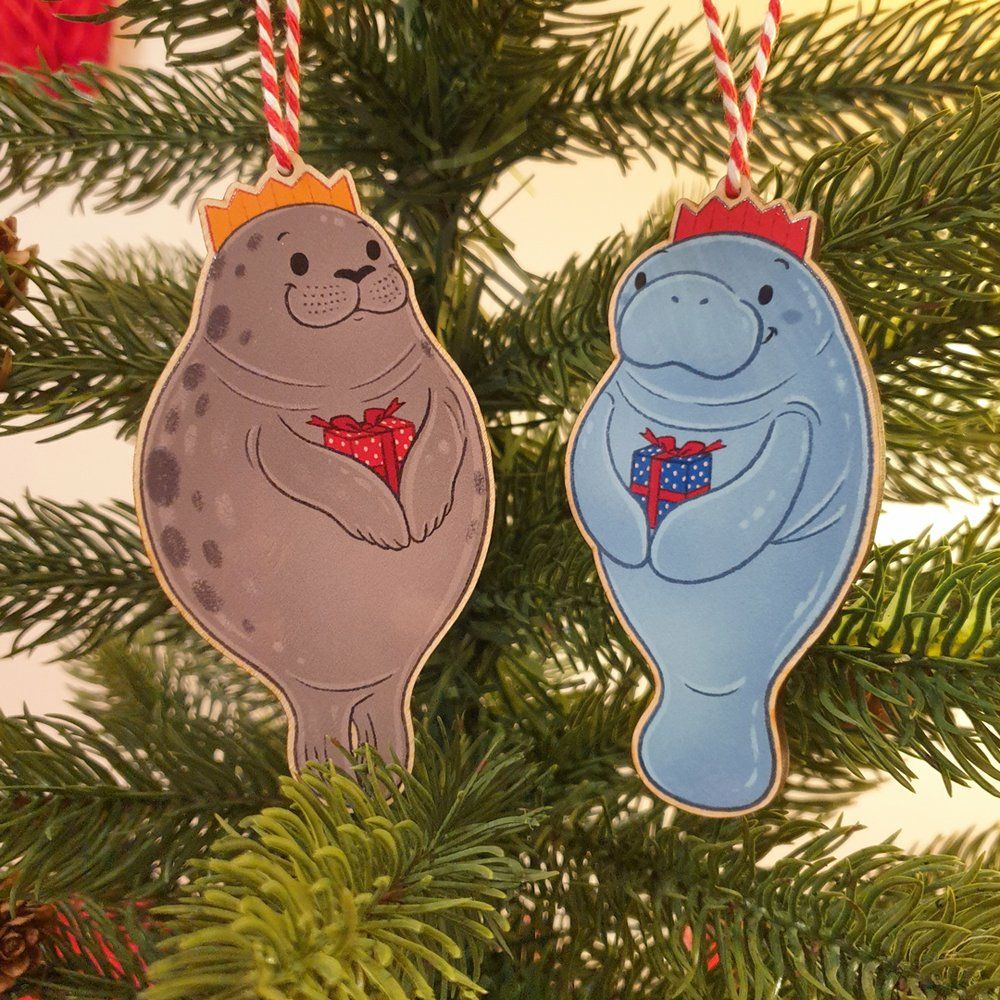 Seal and manatee wooden decorations hanging on a christmas tree. They are smiling and wearing christmas hats and holding a present for each other