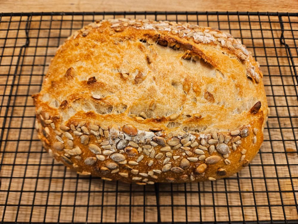 Photo d'une miche de pain refroidissant sur une grille. Il y a des graines de citrouille et de tournesol dessus.