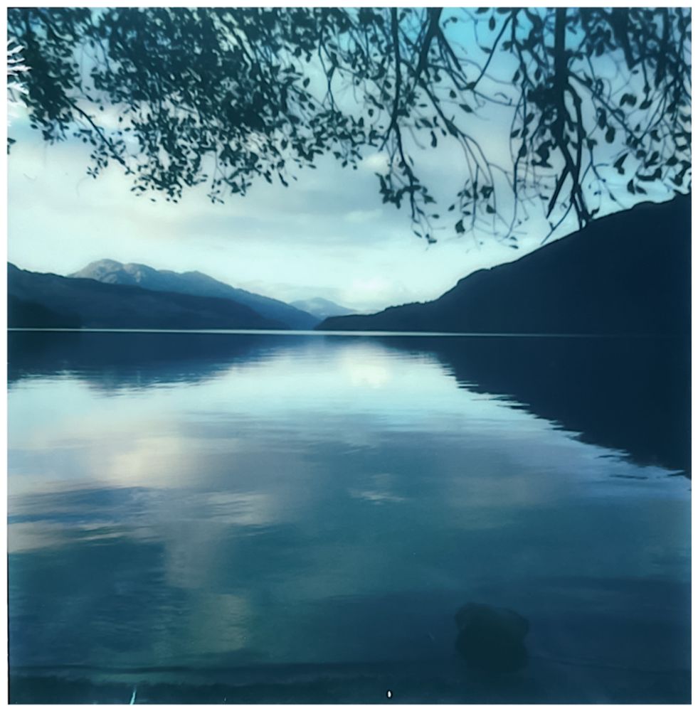 Polaroid image of Loch Lomond taken at Firkin Point. The water is as smooth as glass and stretches towards distant hills. They appear layered against the sky in this low light. Blue, pewter, silver, black and the merest hint of soft purple colour the scene.