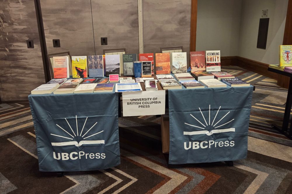 A table in a conference centre. The table features books and brochures. Two blue banners with the UBC Press logo hang from the front of the table.