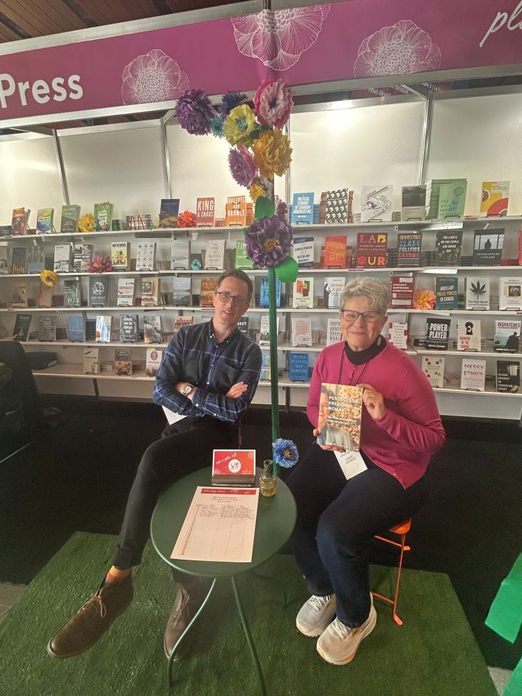 A conference booth with two people sitting on orange chairs around a green table. One, a woman in a pink sweater, holds a copy of a book toward the camera.