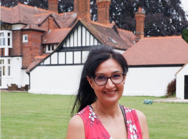 Woman standing and smiling casually in front of a large, brown and white house. 