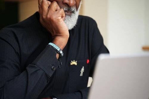 Close-up of an individual using a laptop, focusing on the face and hand. The person is wearing a black shirt with pins, including a crown and a red cross.
