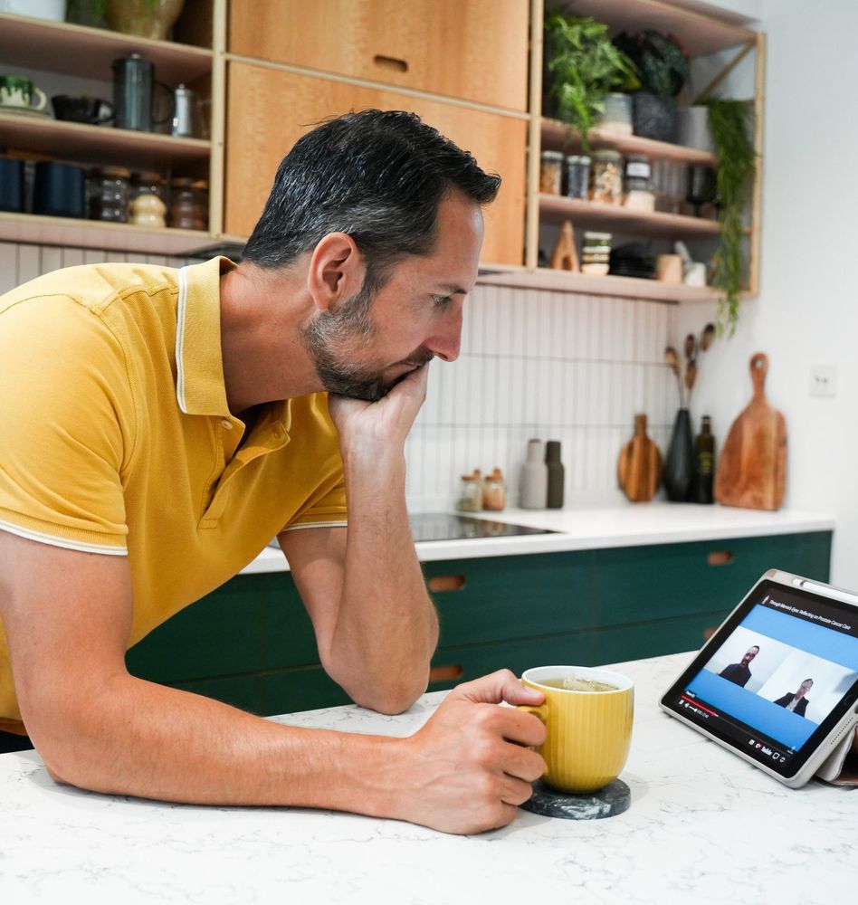 An individual is sitting at a kitchen counter watching a webinar on a tablet and holding a yellow mug. The kitchen has wooden shelves stocked with various kitchenware.