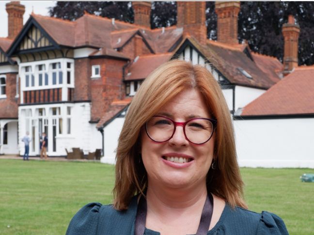 Woman standing and smiling casually in front of a large, white and brown house. 
