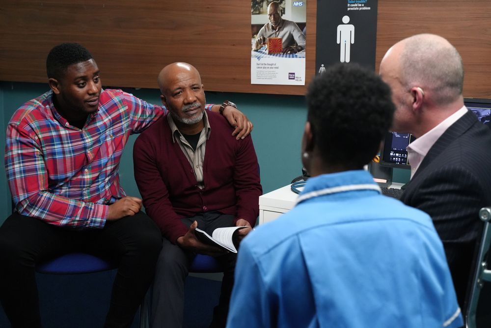 Four people in a discussion inside an office setting. Two individuals are seated and listening intently to another who is speaking, while a fourth observes. A poster reading "NHS" is visible in the background, indicating a healthcare context.