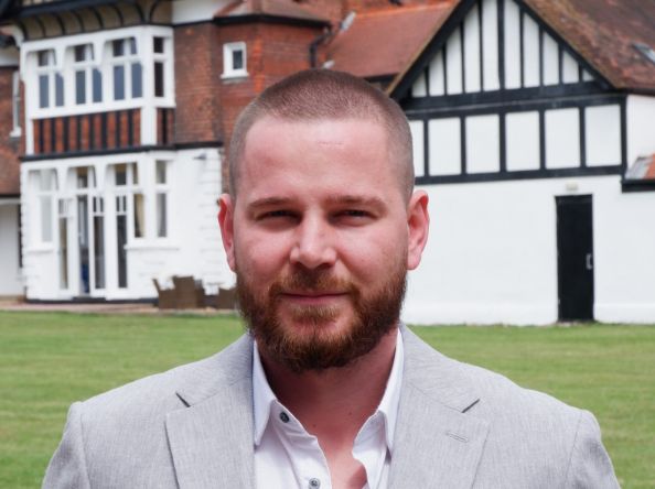 Man in a grey suit standing and smiling casually in front of a house. 