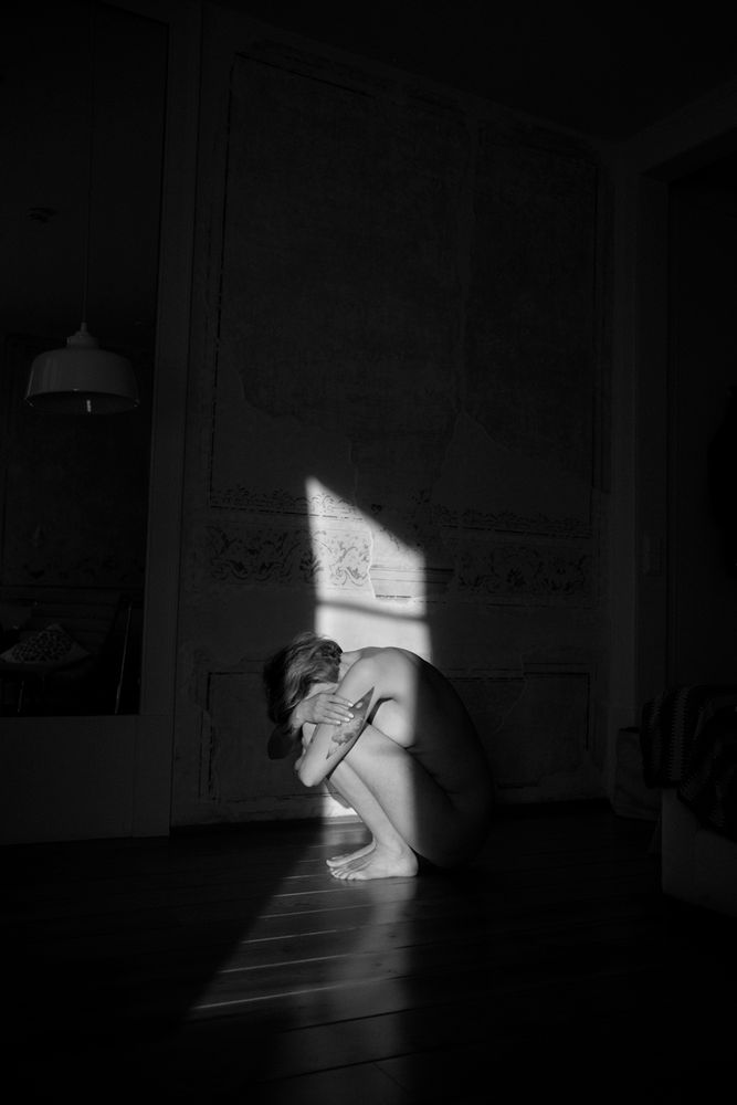 Schwarz weiß Foto. Model hockt seitlich im Lichtschein, der durch die Balkontür auf die Zimmerwand fällt.