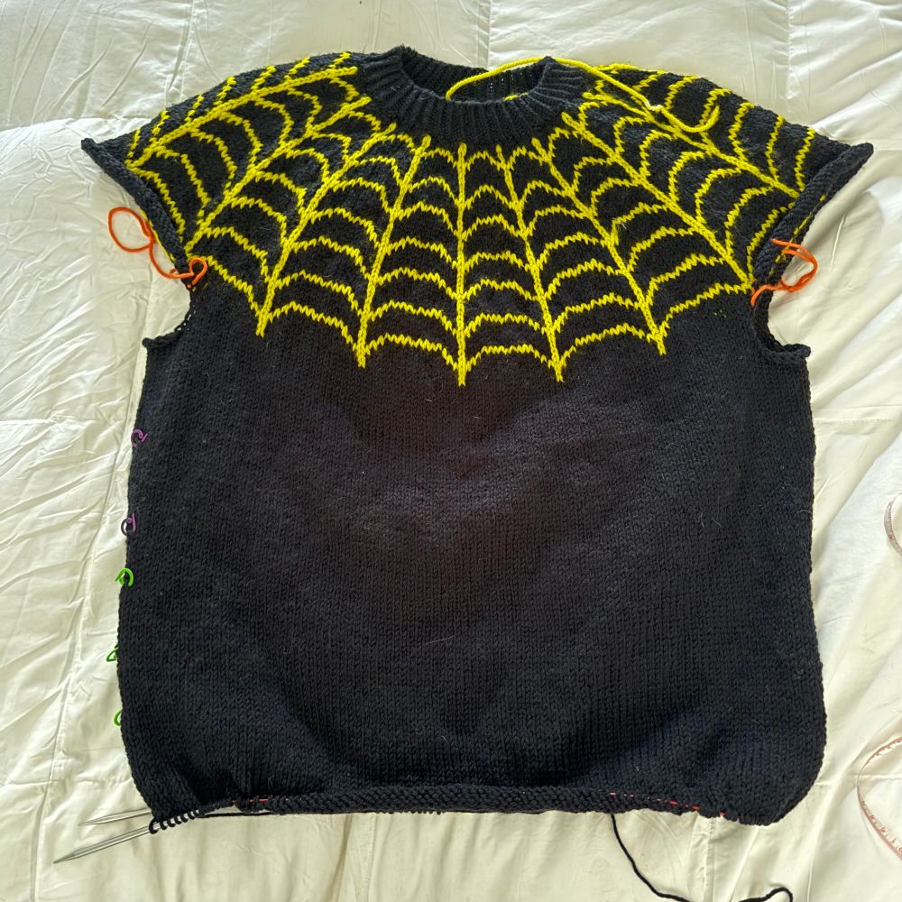 Black knit sweater with a yellow spiderweb motif. The sweater is in progress, and has no sleeves yet. 