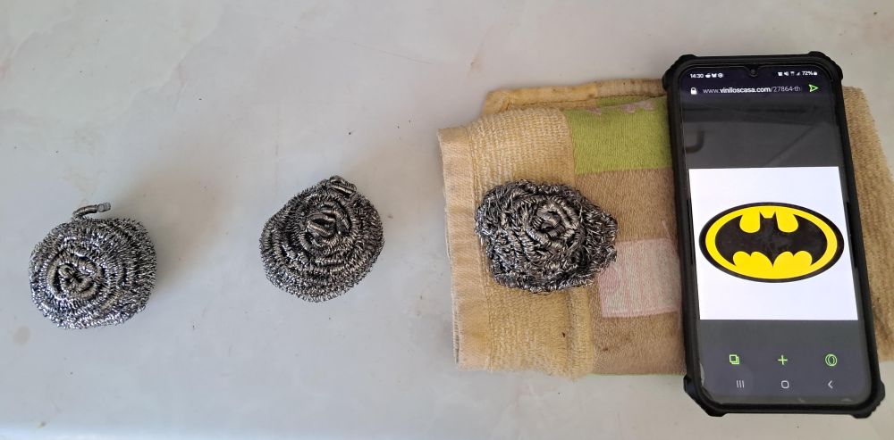 Una foto de mi encimera. Hay tres estropajos nanas puestos en fila, y al lado un móvil con el símbolo de Batman.