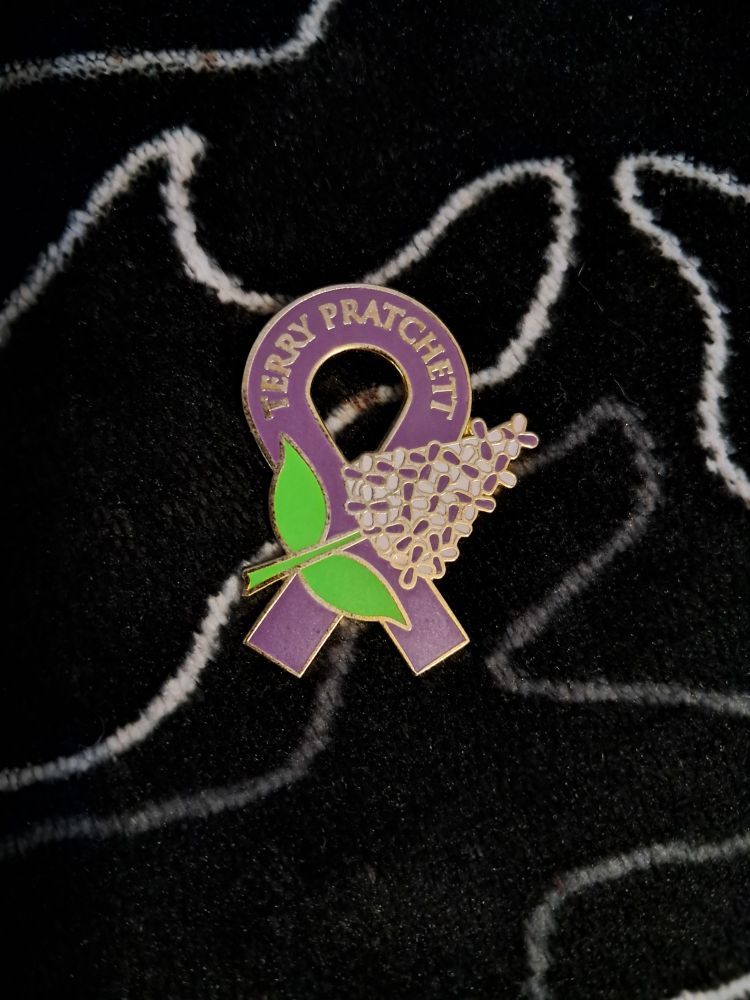 Un pin con un lazo morado y una flor de lila. En lo alto del lazo está escrito TERRY PRATCHETT.