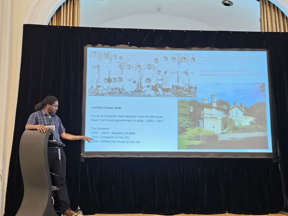 Prof. Sobers presenting slides on a stage. Slides depicting Fairfield House in Bath