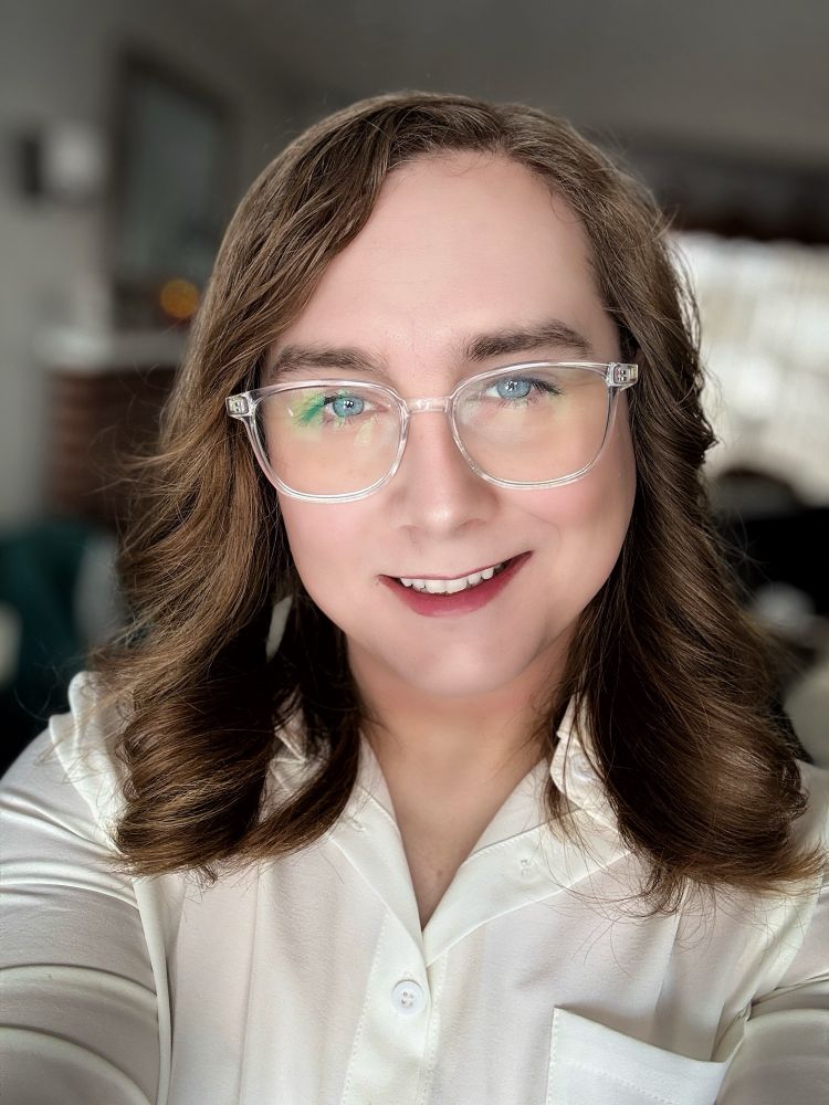 A warm, friendly selfie of Summer with shoulder-length brown hair with subtle highlights.

They’re wearing clear-framed glasses and have bright blue eyes.

Their smile is genuine and welcoming, showing their teeth.

They’re dressed in a white button-up shirt and appear to be sitting in a comfortable indoor setting, with a cozy living space visible in the soft-focused background that includes what looks like furniture and home decor.

The lighting is natural and flattering, creating a relaxed, approachable atmosphere in the photo.​​​​​​​​​​​​​​​​