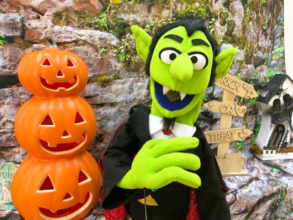 Baron Erdbeere, a green puppet with fangs and black hair, dressed like Count Dracula, stood next to to three stacked Jack O'Lanterns in front of a stone wall with vegetation growing out of it. Behind him is a signpost saying Trick or Treat and at the edge of the photo is a crazy looking little haunted house.