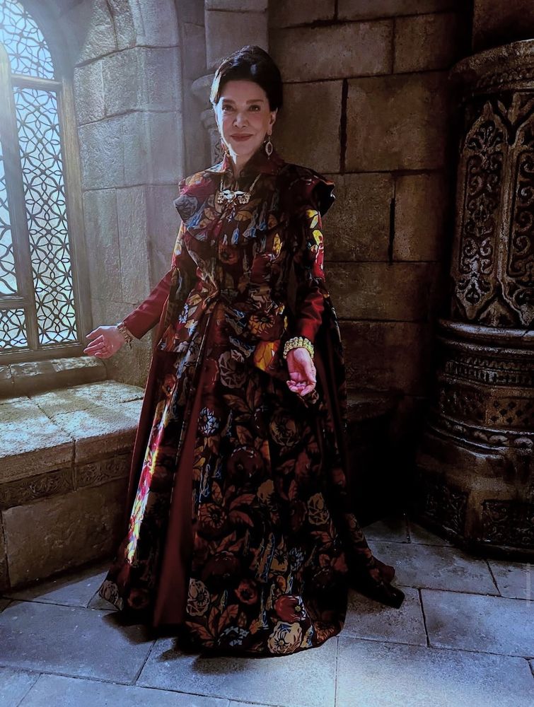 Shohreh Aghdashloo as Elaida in a white tower hallway wearing an ornate floral red dress with an updo
