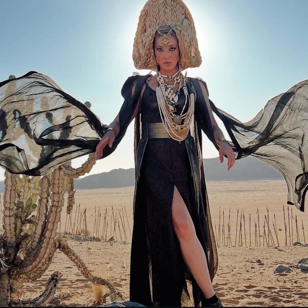 Natasha Culzac as Sevannah in the wheel of time. She’s in the desert, with a tan, braided headdress, a huge cluster of pearl and other necklaces, a black dress belted with gold, and long filmy sleeves. Her sleeves are fanned out around her in the picture as if tossed into the air for the shot.