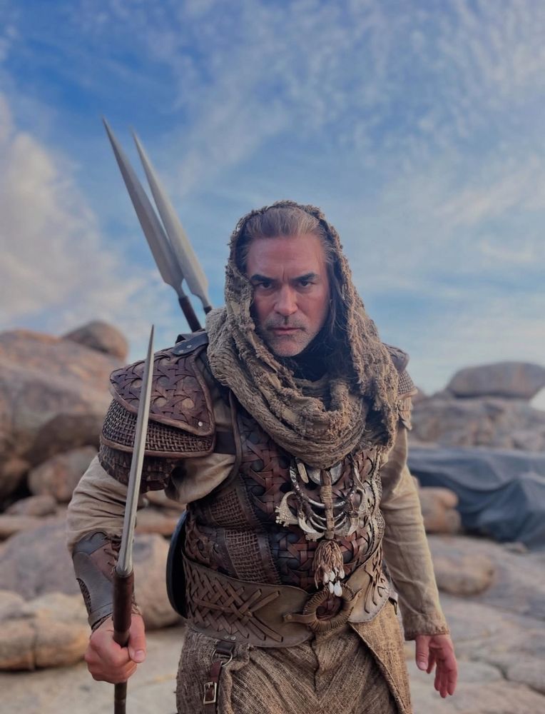 Bjoern Landberg as Rhuarc is Aiel garb complete with spears on his back and in his hand, a shoufa, and intricate necklace passed down from chief to chief in Rhuarc’s clan